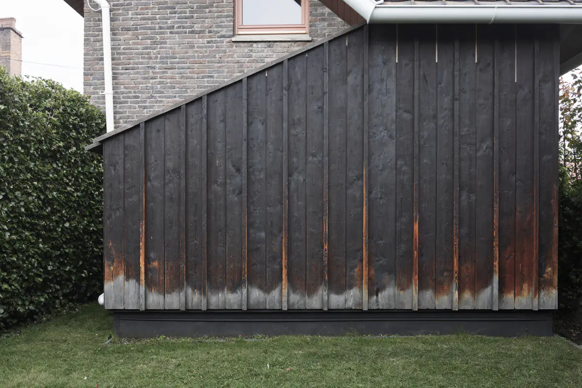 Verticale donker gebeitste houten gevelbekleding van een aanbouw naast een bakstenen gevel, onderaan verweerd