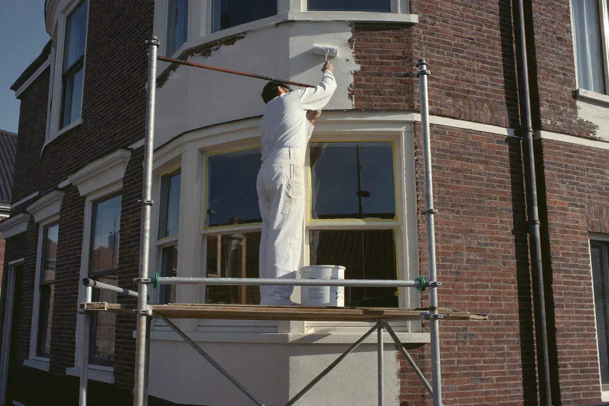 Schilder op een steiger verft een wit geschilderde bakstenen gevel van een hoekhuis